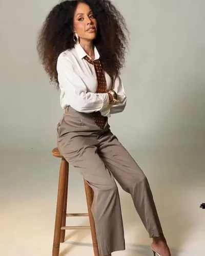 Mulher de cabelo cacheado, camisa branca, gravata e calça cinza, sentada num banco alto de madeira em fundo branco