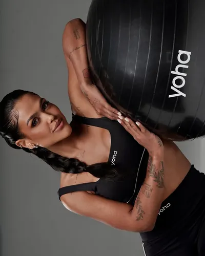 Camila Loures de top preto segurando uma bola de pilates da marca Yoha, em fundo cinza